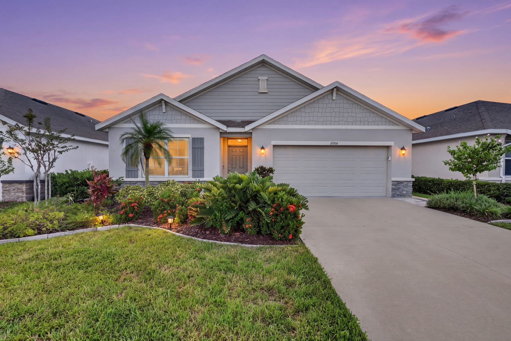 Front of the gray craftsman-style home at sunset with lit coach lights, a palm tree and tropical landscaping