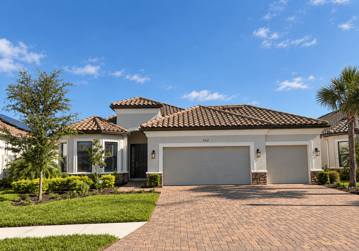 A single-story Mediterranean-style home with a clay tile roof, cream stucco, stone accents, a three-car garage and a paver driveway under a blue sky