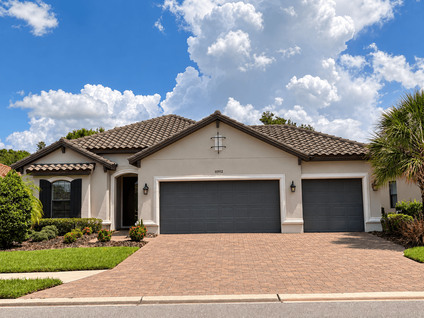 A single-story Mediterranean-style home with a brown tile roof, beige stucco, dark shutters, charcoal garage doors and a paver driveway under a blue sky with tall white clouds