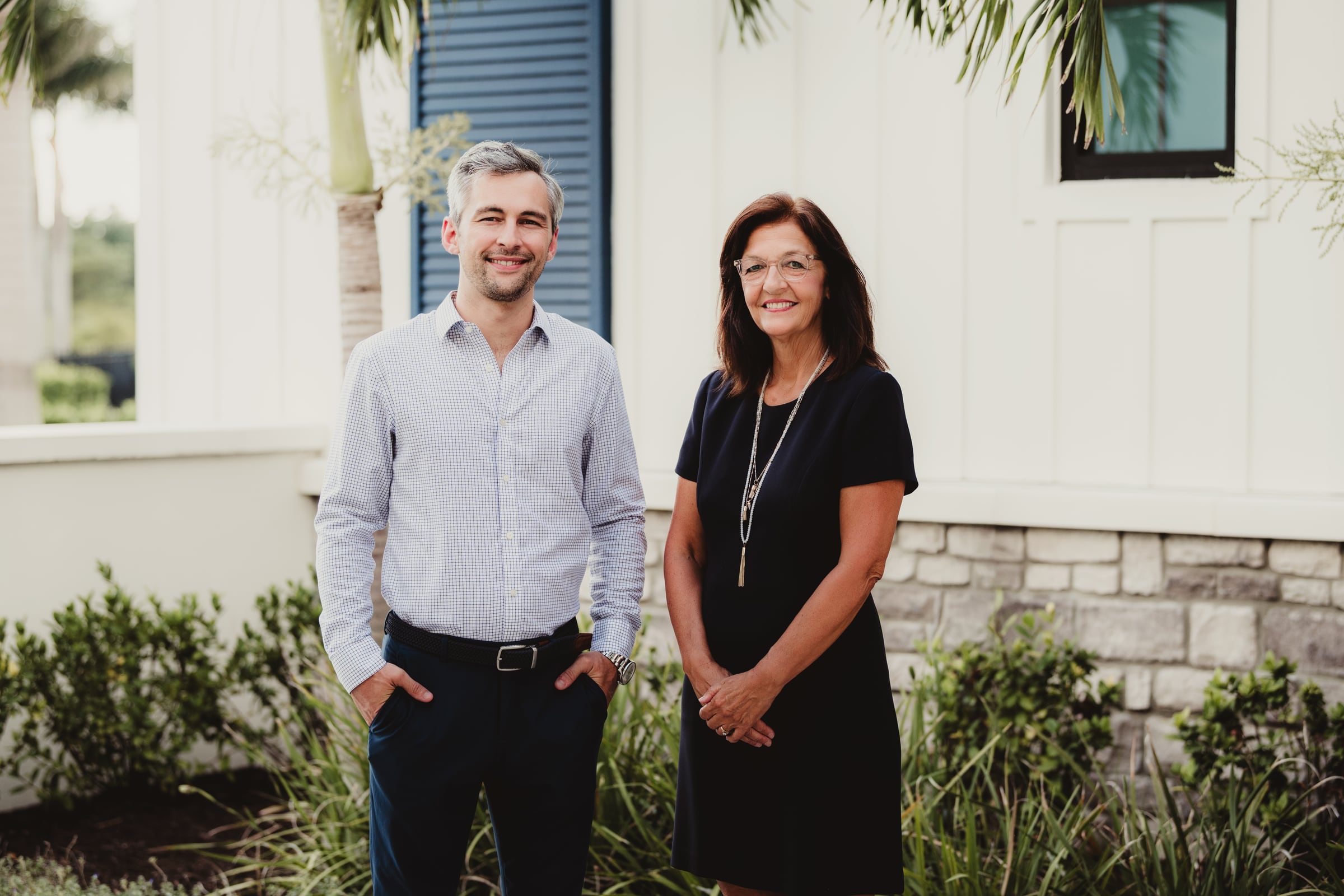 Bryan and Tammy Sinkel standing together outside a white home with blue shutters and palm trees